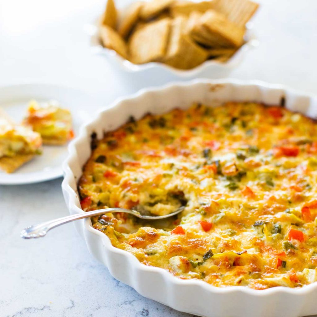 A white dish of baked jalapeno artichoke dip has a spoon serving a bit next to a bowl of crackers.