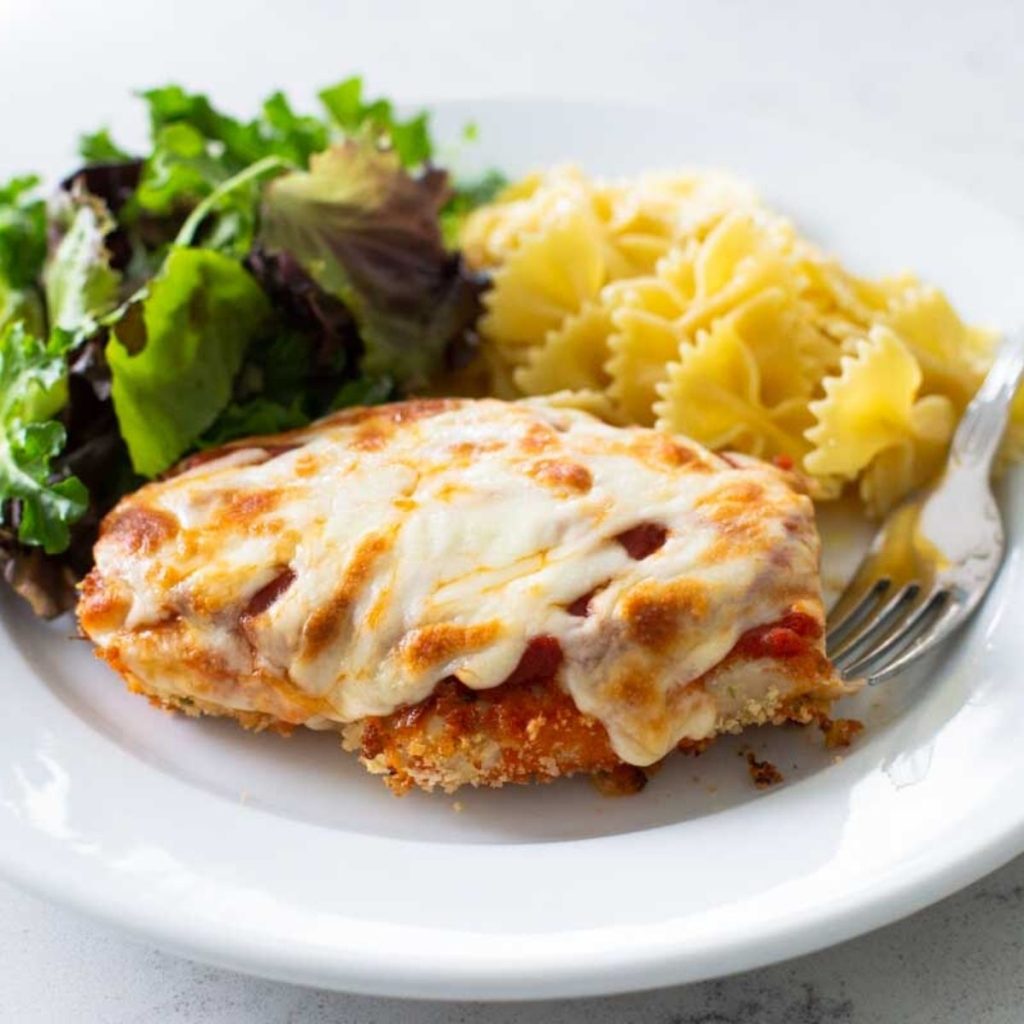 A breaded chicken cutlet topped with tomato sauce and melted cheese is served with a green salad and some bowtie pasta on the side.