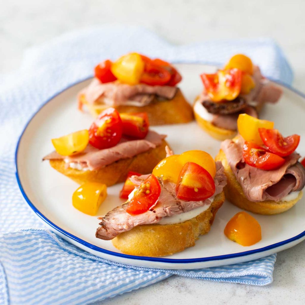 A plate of crostini toasts topped with roast beef and cherry tomatoes.