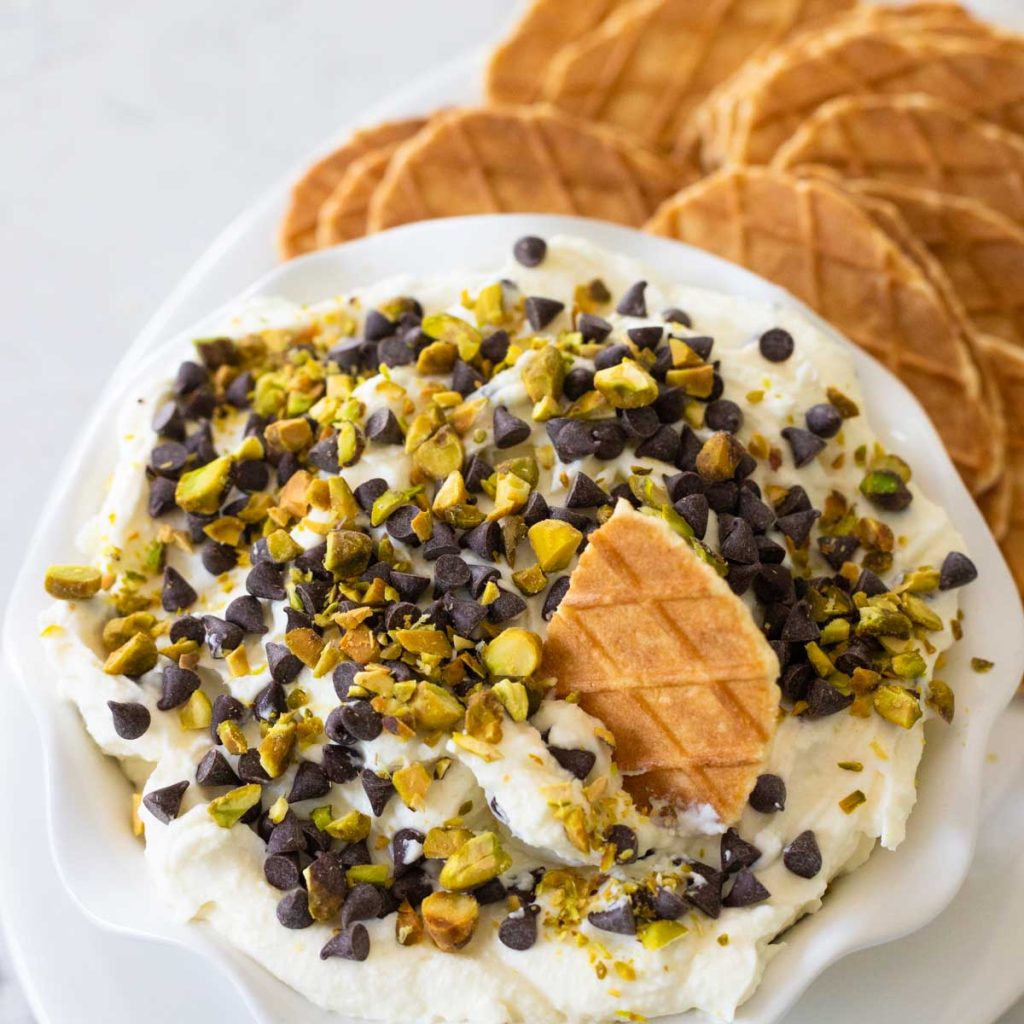 A bowl of creamy cannoli dip has chopped pisatchios and chocolate chips on top. Waffle cookies are served on the side.