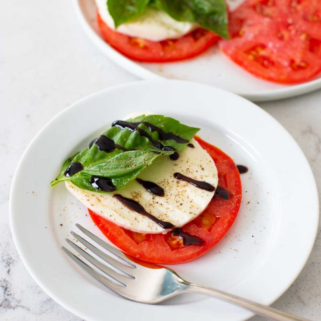 A sliced tomato, mozzarella, and fresh basil appetizer with a balsamic drizzle is on a white plate.