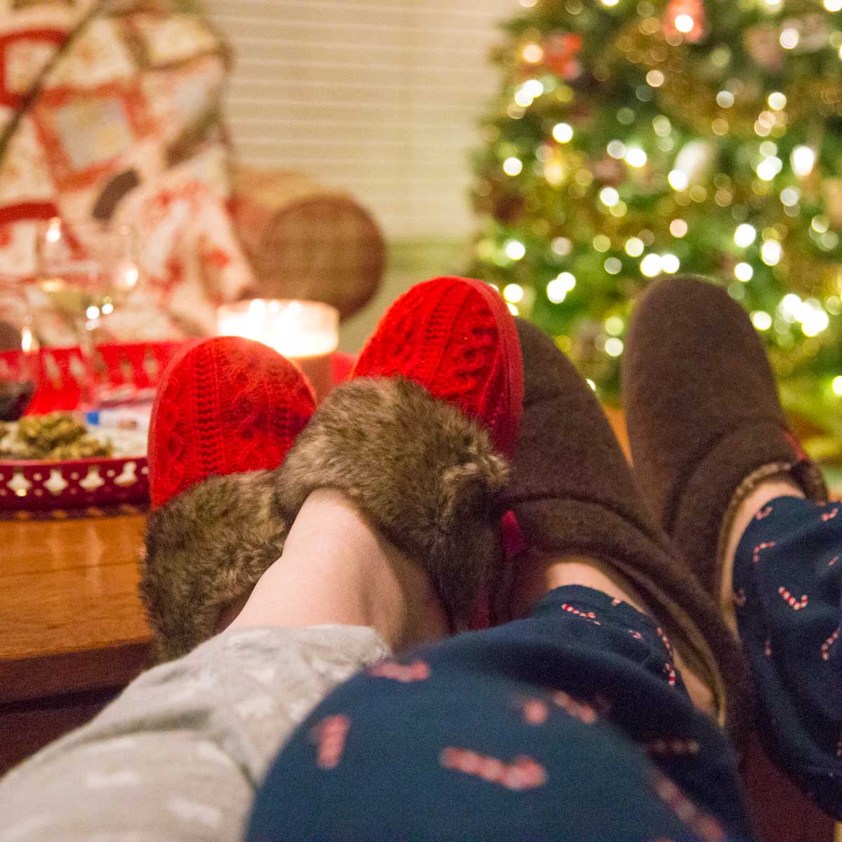 A husband and wife wearing cozy slippers snuggle by the Christmas tree.