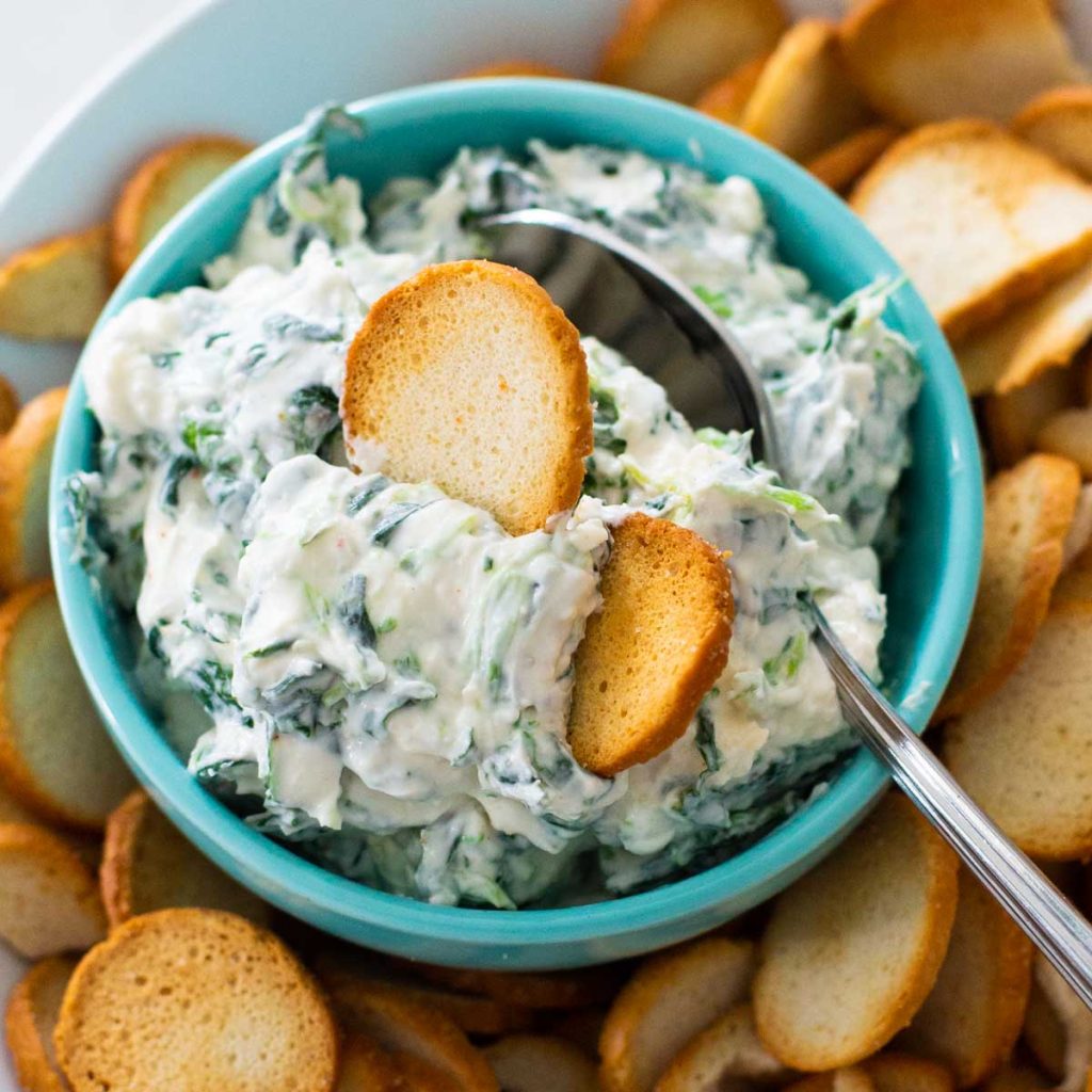 A blue bowl is filled with spinach dip, there are bagel chips all around.