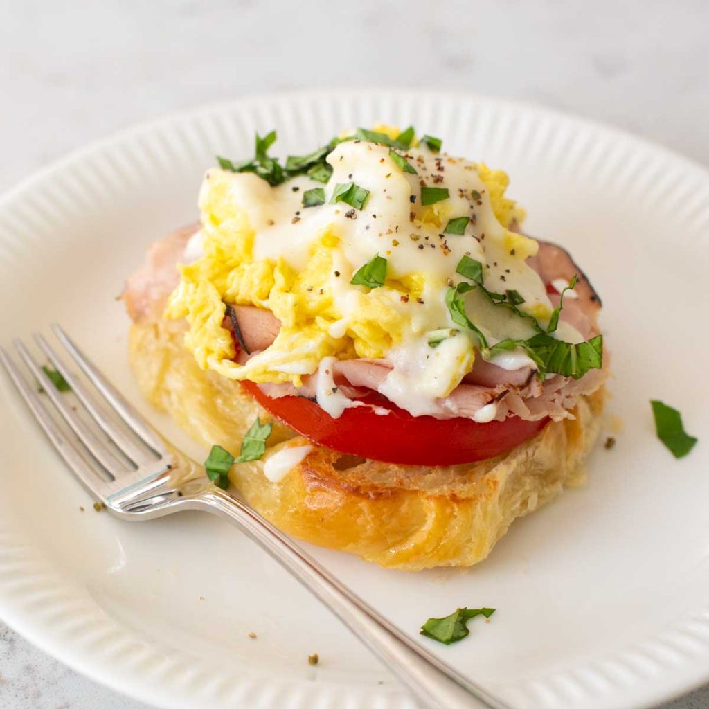 A scrambled eggs benedict on a croissant with fresh tomato and a cream sauce drizzled over the top.