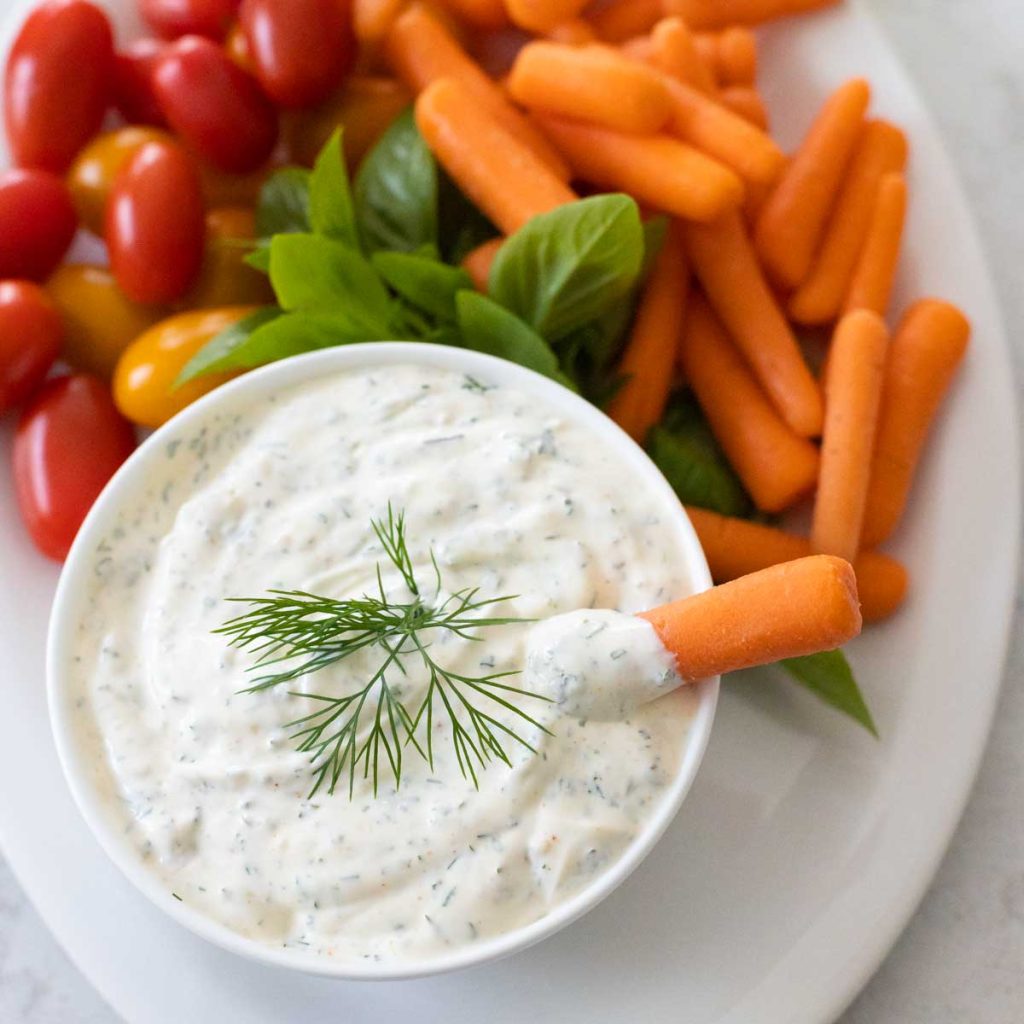 a bowl of homemade dill dip has baby carrots and cherry tomatoes on the plate next to it.
