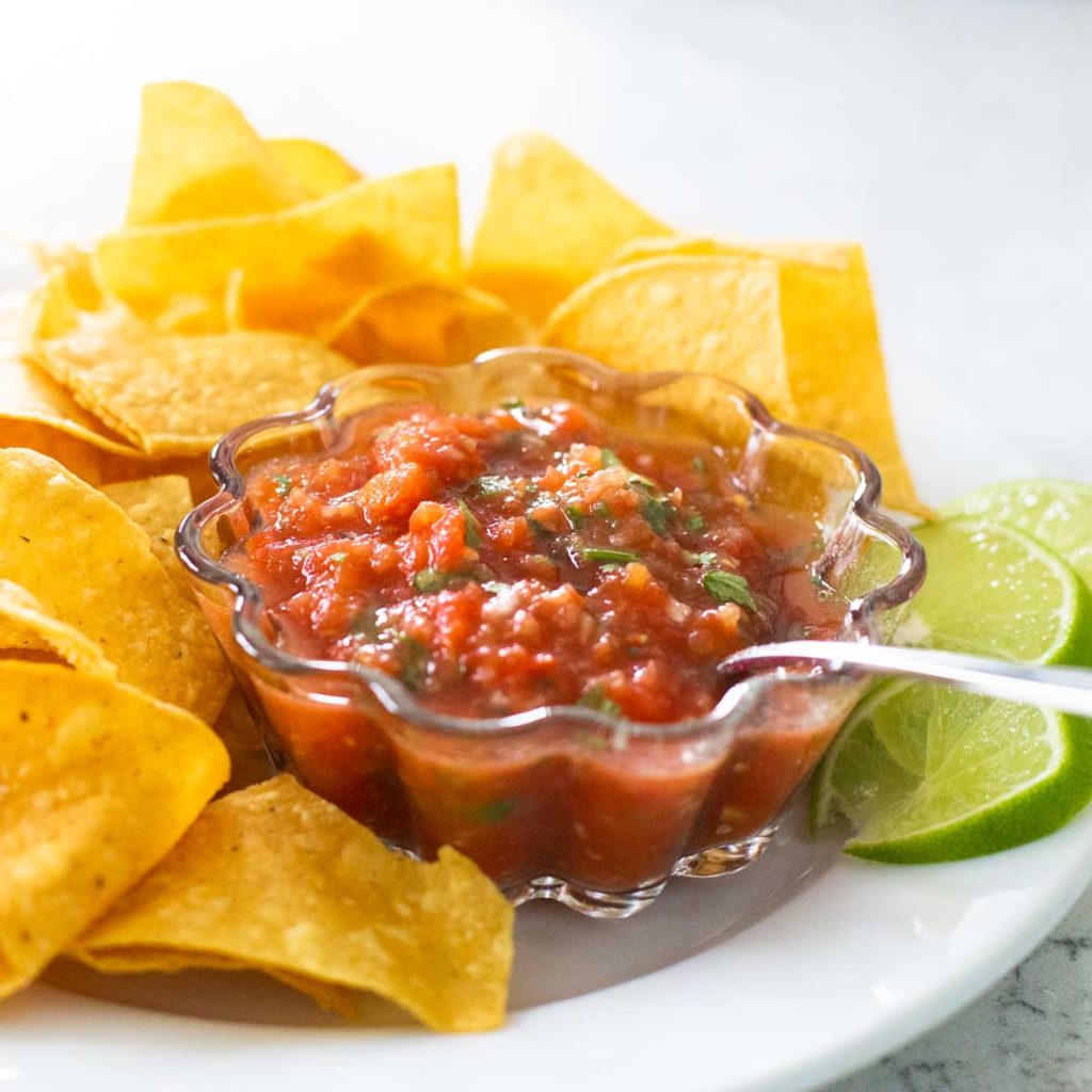 A bowl of fresh salsa and tortilla chips with fresh lime wedges on the side.