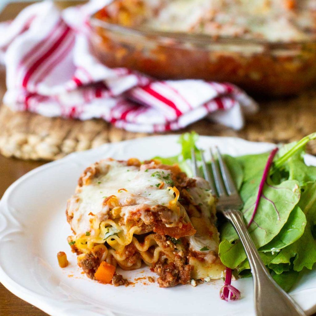 A serving of lasagna with ruffled edges is served on a white plate with a spinach salad.