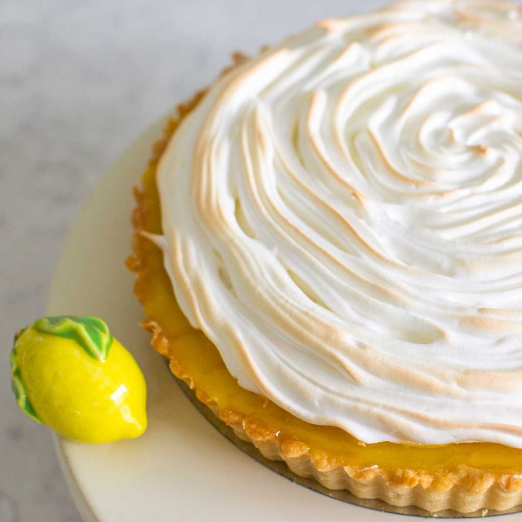 A lemon tart with meringue topping is on a cake plate with a ceramic lemon ornament.