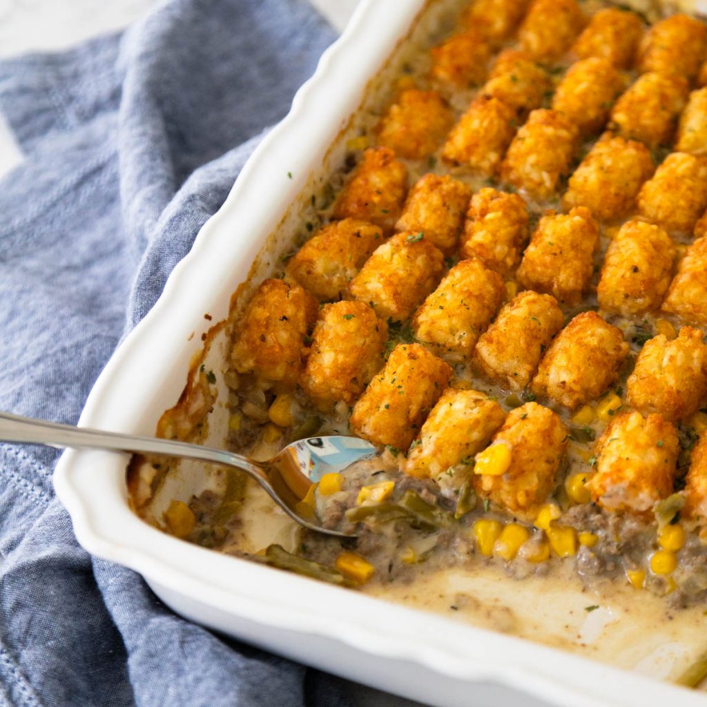 A white baking dish has a tater tot topped casserole with a spoon serving a scoop.