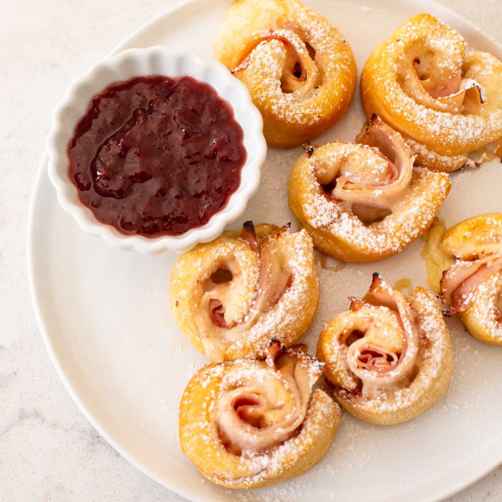 a white platter has several crescent roll pinwheels next to a bowl of jam.