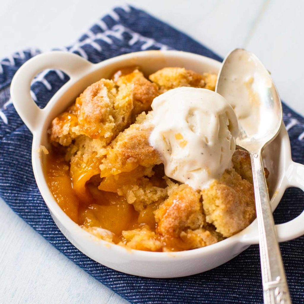 A white dish has a serving of fresh peach cobbler with a scoop of vanilla ice cream.