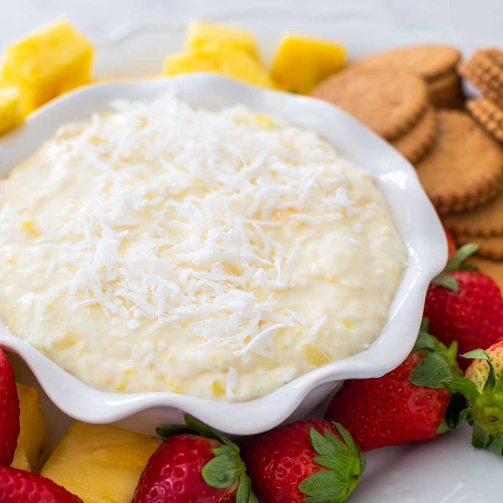 A bowl of creamy dip has fresh strawberries, pineapple, and ginger cookies on the plate next to it.