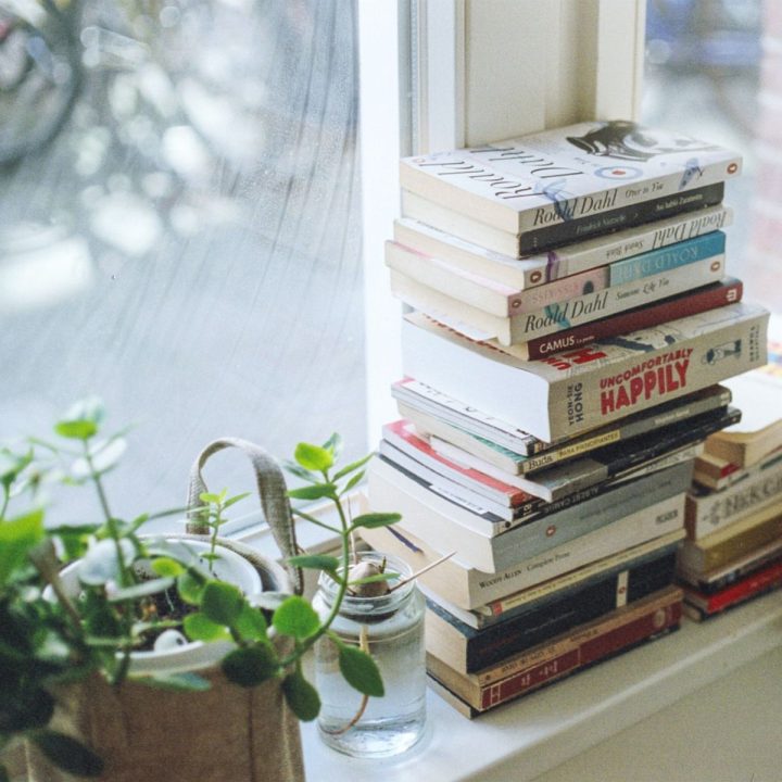 A stack of books sits in a window sill next to a green plant.