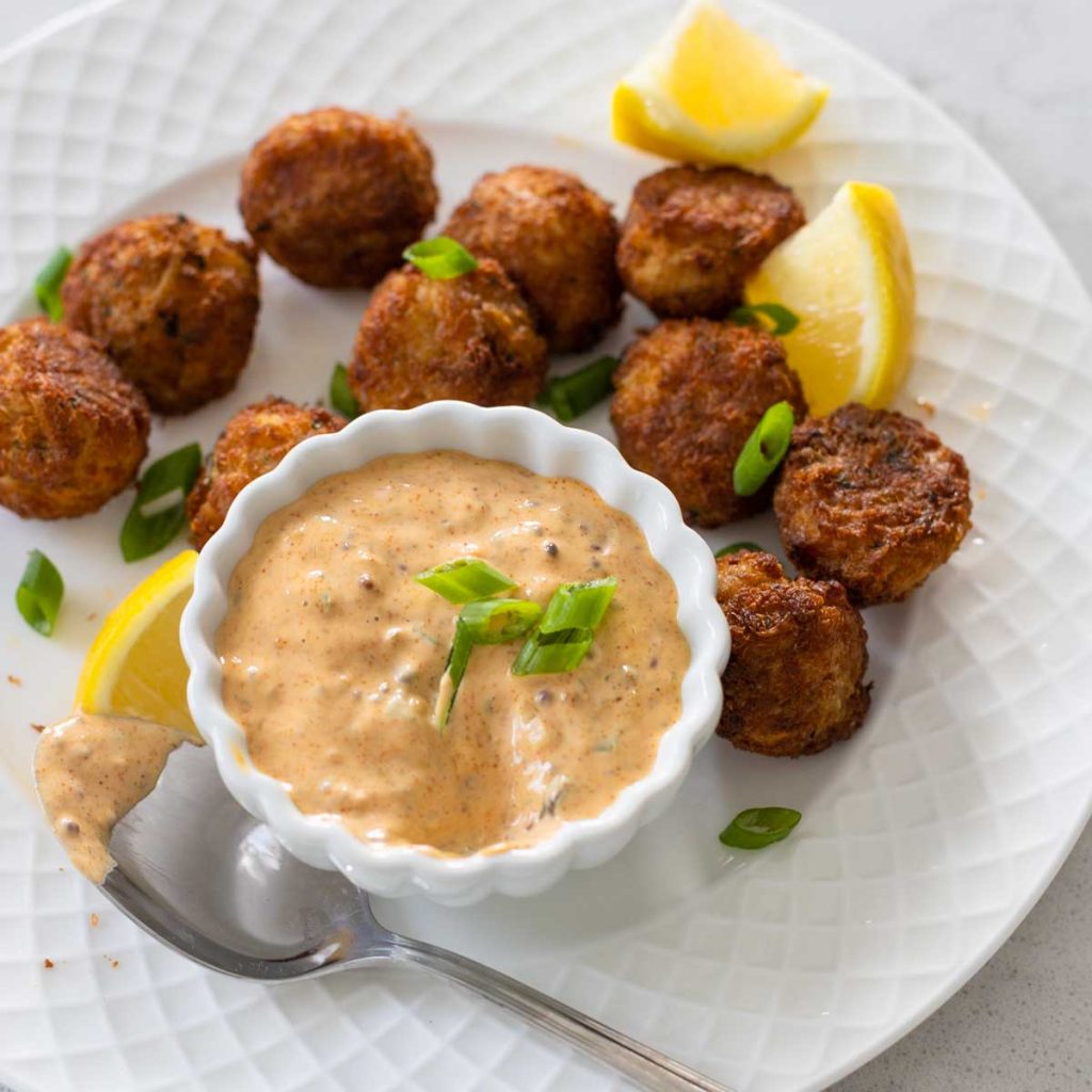 A plate of mini crab cakes with a cup of remoulade sauce. Fresh lemons are scattered about.