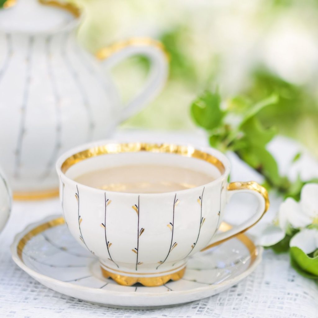 An elegant white tea cup and sauce has gold detailing and sits in front of green floral sprig.