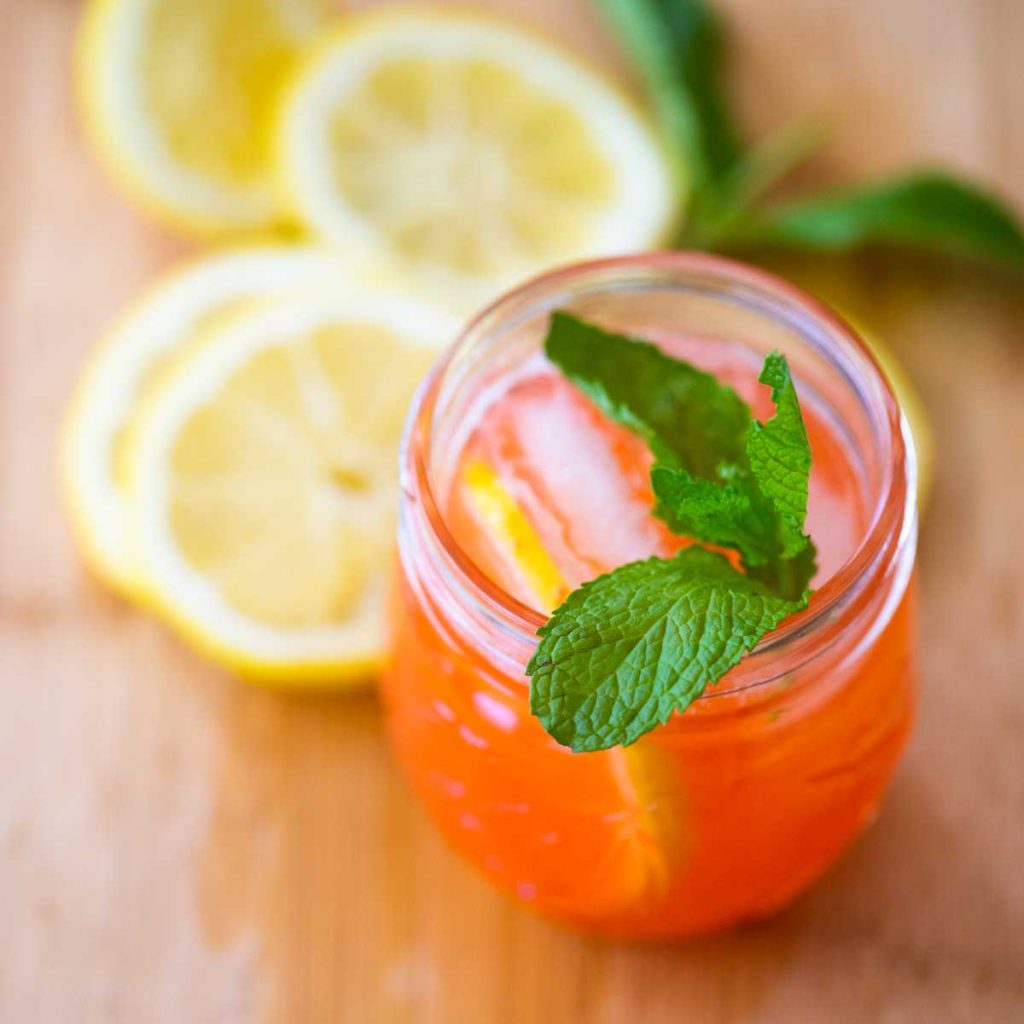 A glass of pink lemonade with fresh mint and sliced lemons in the background.