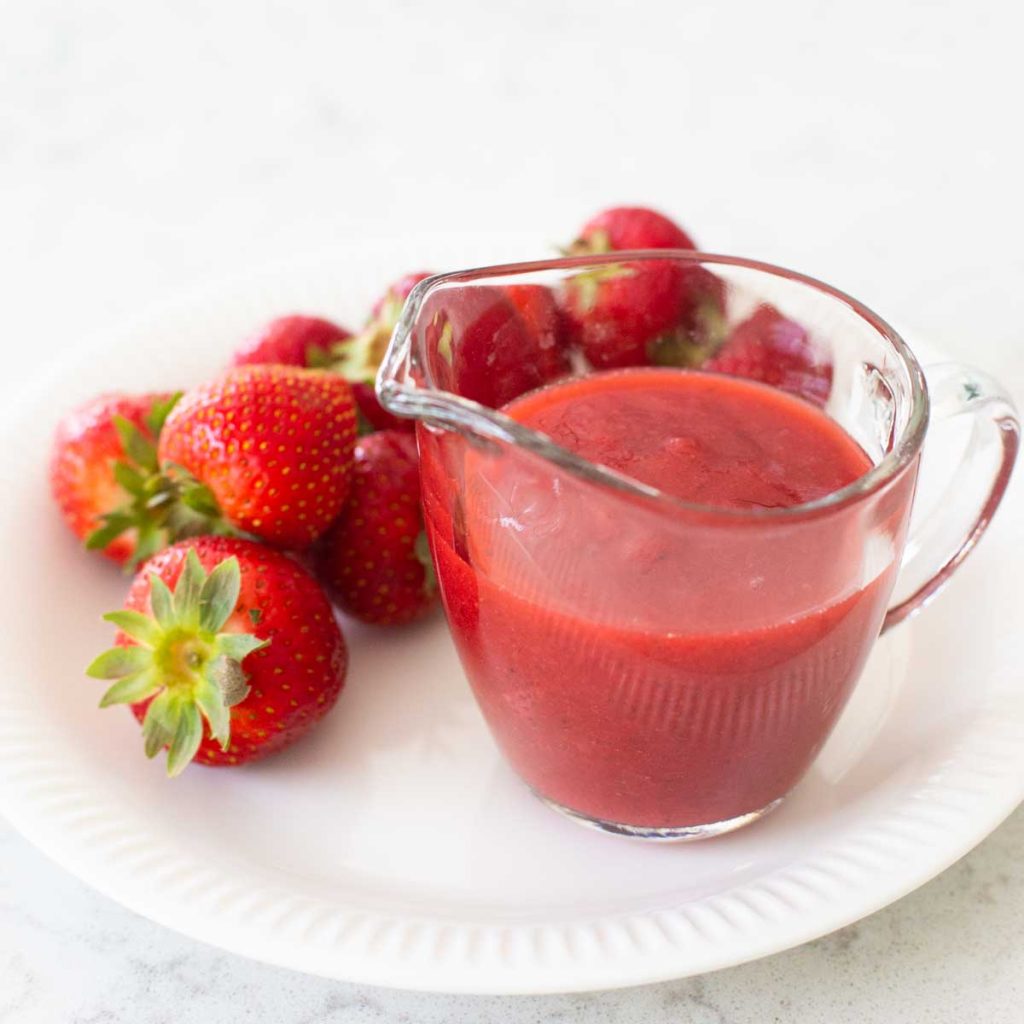 A pitcher of red strawberry sauce sits on a plate next to fresh strawberries.