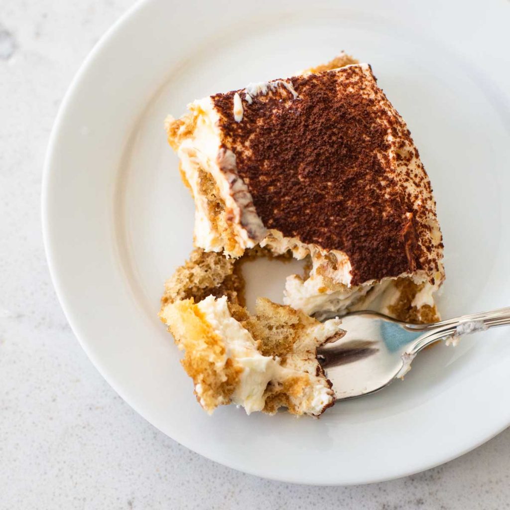 A square serving of tiramisu is on a white plate with a spoon.