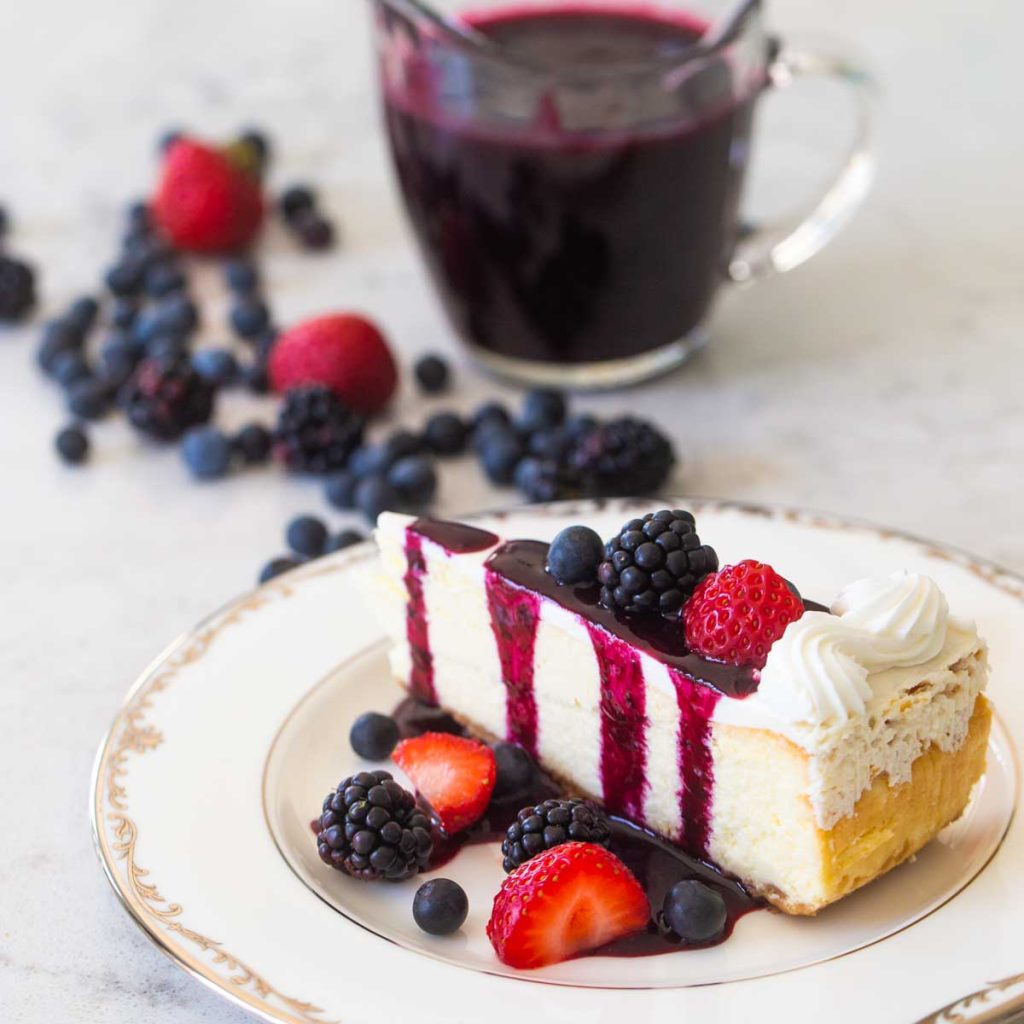 A vanilla cheesecake slice has a drizzle of berry sauce and fresh berries on the plate