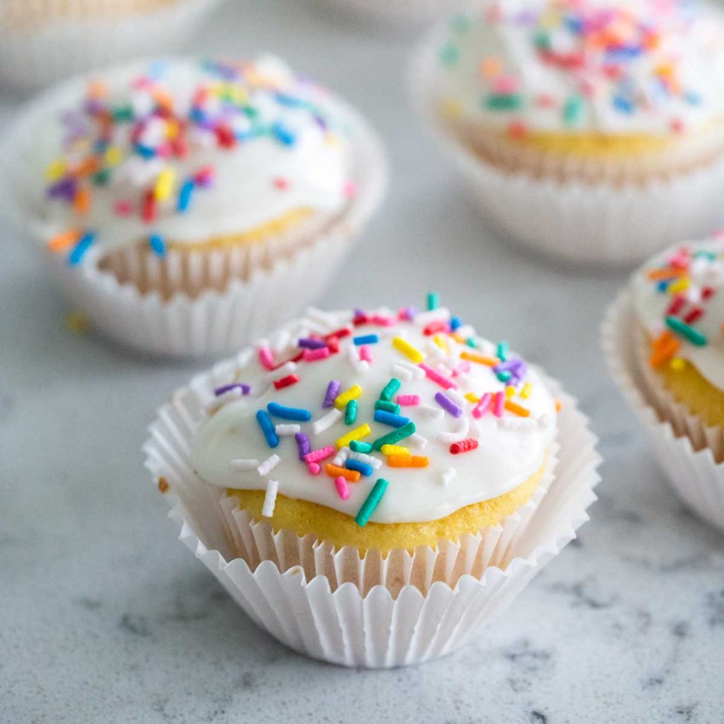 A row of vanilla cupcakes with white icing and sprinkles on top.