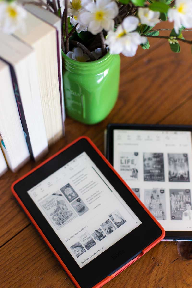 Two Kindle devices are on a table next to a stack of books and a vase of flowers.