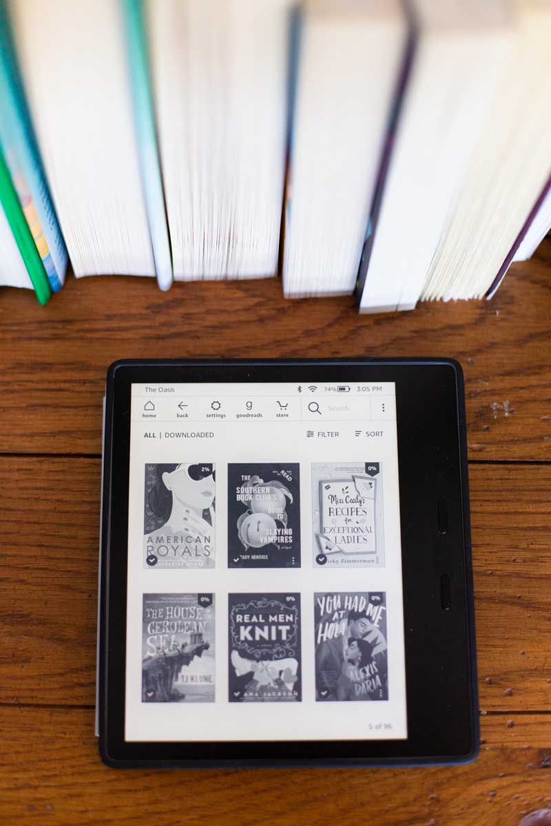A Kindle Oasis sits on a table next to a stack of paper books.