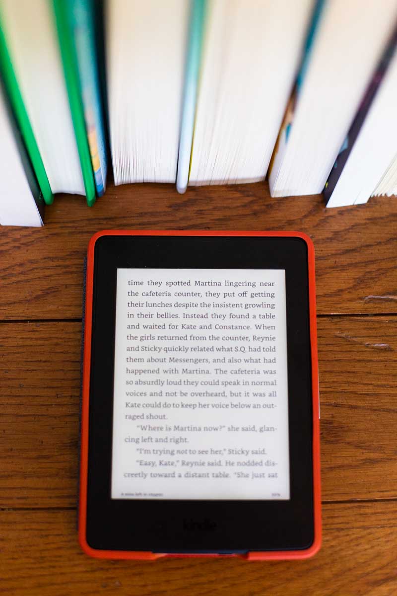 A Kindle Paperwhite sits on a table next to a stack of books.