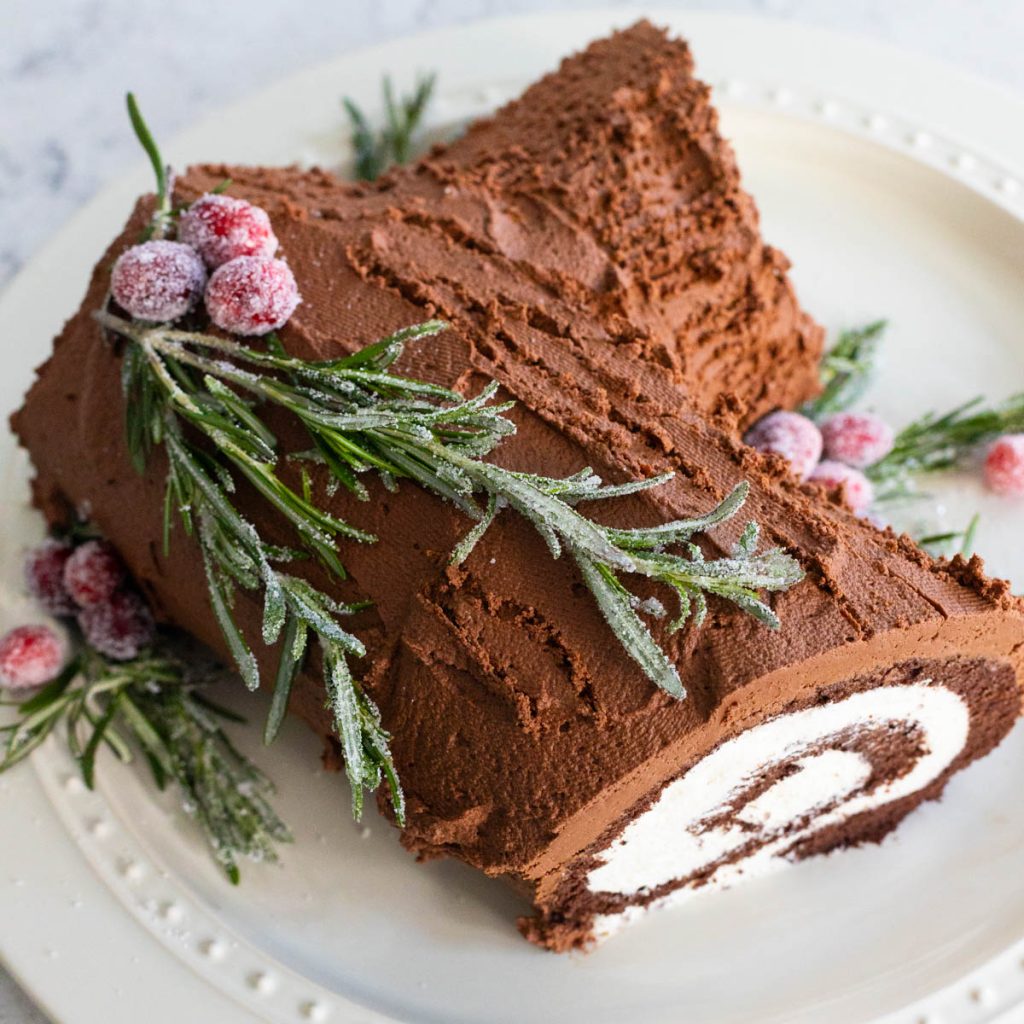 A homemade yule log cake has chocolate frosting and sugared rosemary and cranberries.