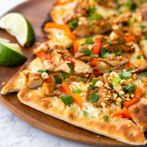 A Thai chicken flatbread has green onions, red peppers, and chopped peanuts on top. Lime wedges on the side.