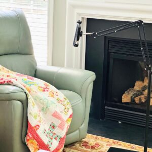 A bendable arm stand rests on the floor in front of a cozy arm chair with a quilt ready for snuggling. The Kindle has been mounted and is ready to read.