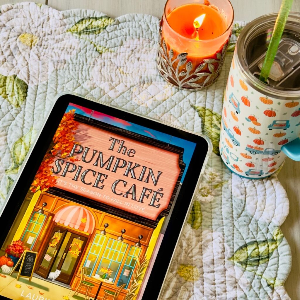 The cover of the book The Pumpkin Spice Cafe by Laurie Gilmore is on the screen of an iPad sitting next to a pumpkin coffee mug and an orange candle.