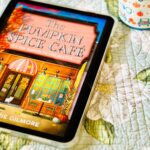 The cover of the book The Pumpkin Spice Cafe by Laurie Gilmore is on the screen of an iPad sitting next to a pumpkin coffee mug and an orange candle.