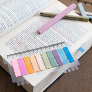 A book is open and has a strip of colorful sticky tabs, pastel highlighters, and a fineline pen ready to annotate the pages.