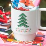 A cute holiday mug stuffed with hot cocoa packets sits on a pile of Christmas books in front of a Christmas tree.