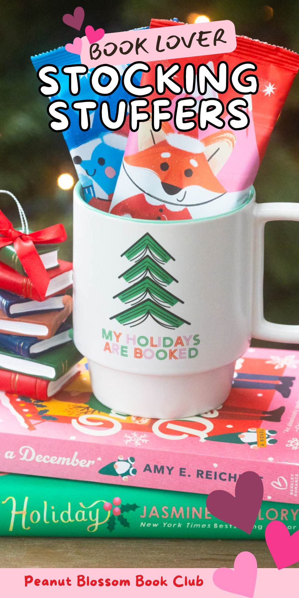 A cute holiday mug stuffed with hot cocoa packets sits on a pile of Christmas books in front of a Christmas tree.