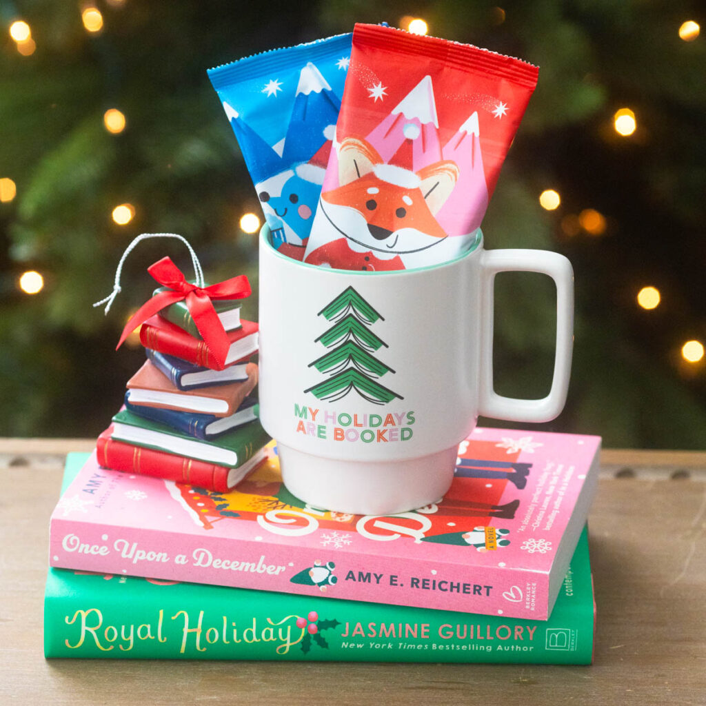 A cute holiday mug stuffed with hot cocoa packets sits on a pile of Christmas books in front of a Christmas tree.