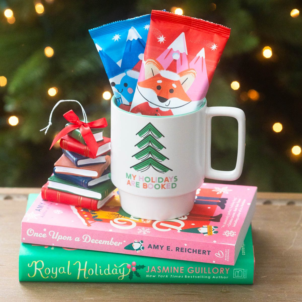 A cute holiday mug stuffed with hot cocoa packets sits on a pile of Christmas books in front of a Christmas tree.