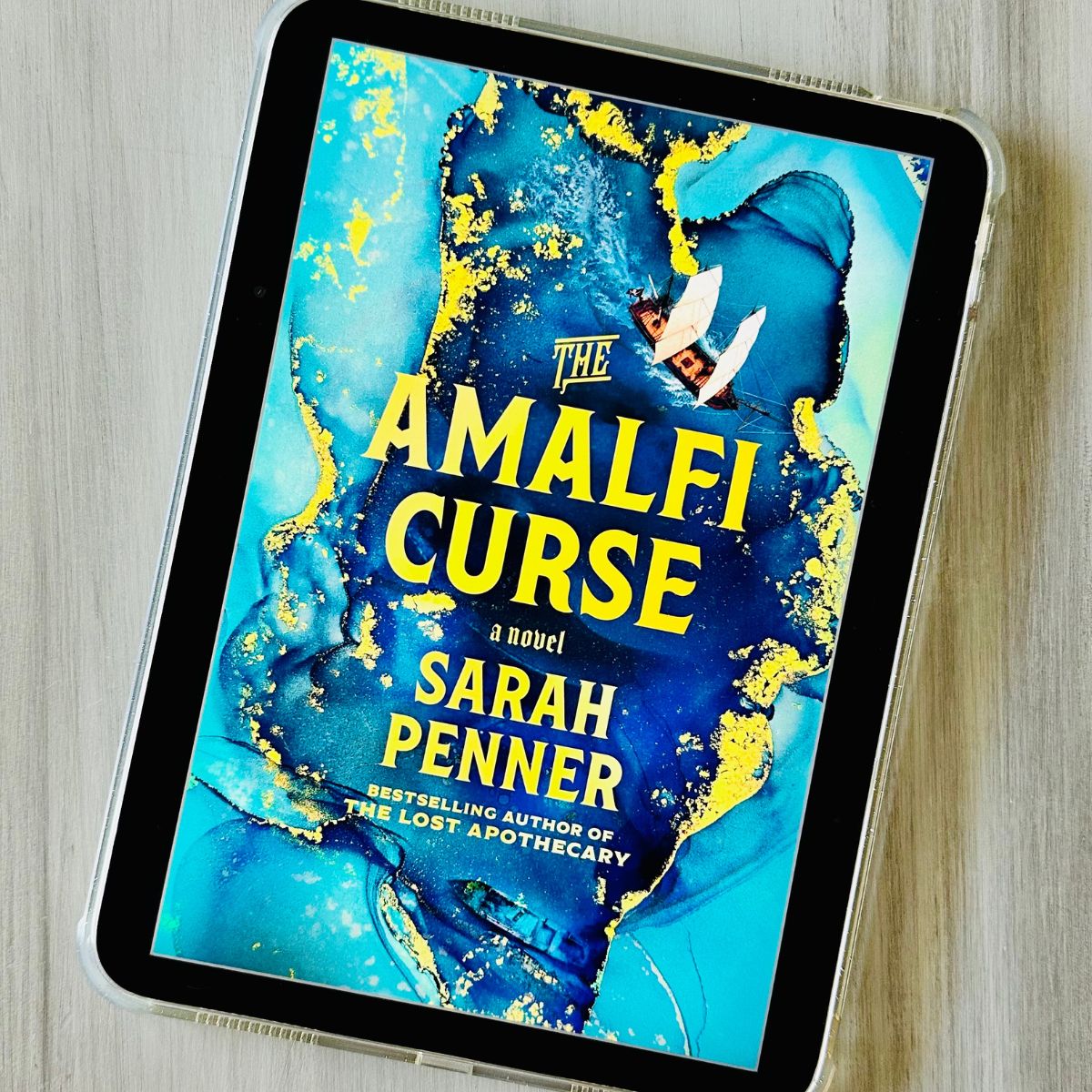 The cover of the book The Amalfi Curse by Sarah Penner is on the screen of an iPad.