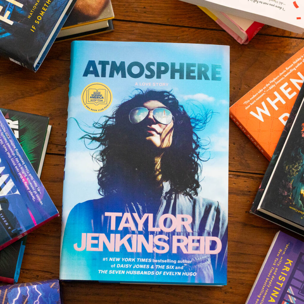 A copy of the book Atmosphere by Taylor Jenkins Reid sits on a table surrounded by more books.